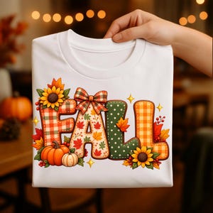 Puede incluir: Una camiseta blanca con la palabra FALL en letras grandes y decorativas. Las letras tienen patrones de cuadros rojos y blancos, hojas de otoño, lunares verdes y vichy naranja. El diseño incluye girasoles, calabazas y un lazo de cuadros.