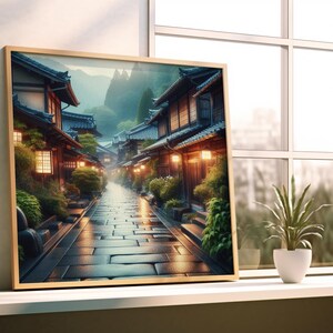 Könnte beinhalten: Ein gerahmter Druck einer japanischen Straßenszene. Das Kunstwerk zeigt eine Kopfsteinpflasterstraße mit traditionellen Holzgebäuden, die von warmem Licht beleuchtet werden. Üppiges Grün und ein nebliger Bergrücken verleihen der Szene Tiefe.