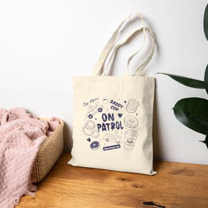 Può includere: Una borsa tote color crema con un design grafico blu navy. Il design include un'auto della polizia, ciambelle e le parole "Cop Moms" e "Daddy Cop ON PATROL". La borsa ha due manici ed è realizzata in tela.