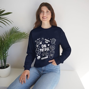 Può includere: Felpa blu navy con illustrazioni e testo bianchi. Il design include le parole "Daddy Cop On Patrol" insieme a immagini di un'auto della polizia, ciambelle e una tazza di caffè. È presente anche l'hashtag #grumpycop.