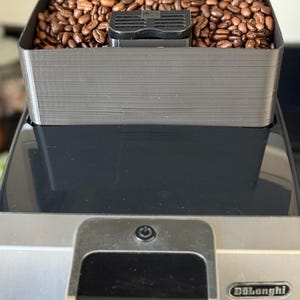 Puede incluir: Primer plano de una cafetera con una tolva de granos llena de granos de café tostados. La máquina tiene un diseño negro y plateado, con la marca "DeLonghi" visible. La pantalla de la máquina muestra texto en francés.