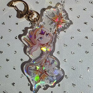 May include: A clear acrylic keyring featuring a cartoon character with pink hair, holding a star-shaped staff. The keyring has a gold-coloured clasp and a holographic effect, creating a shimmering appearance.
