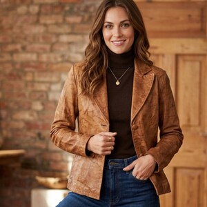 May include: A brown leather blazer with a classic design. The blazer has a notched lapel, two buttons, and a textured finish. The model is wearing a brown turtleneck and blue jeans. A gold necklace is visible.