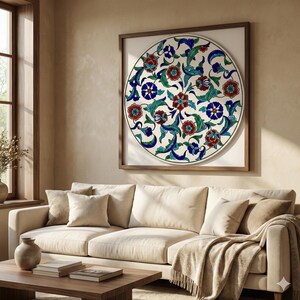 May include: A framed, circular artwork featuring a floral design in red, blue, and green on a white background. The artwork is displayed above a cream-colored sofa in a living room setting. The frame is a dark brown color.
