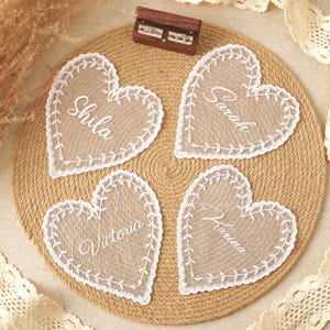 May include: Four heart-shaped beige fabric coasters with white lace trim and embroidered names Shila, Sarah, Victoria, and Hanna. A small rectangular wooden box containing two silver rings sits at the top centre of the circular jute mat.