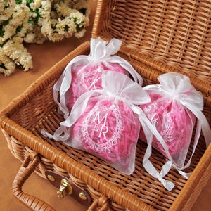 May include: Three sheer white organza drawstring bags filled with bright pink shredded paper filler. Each bag features a white printed monogram design with ornate floral borders. The bags are arranged inside a woven brown wicker basket.