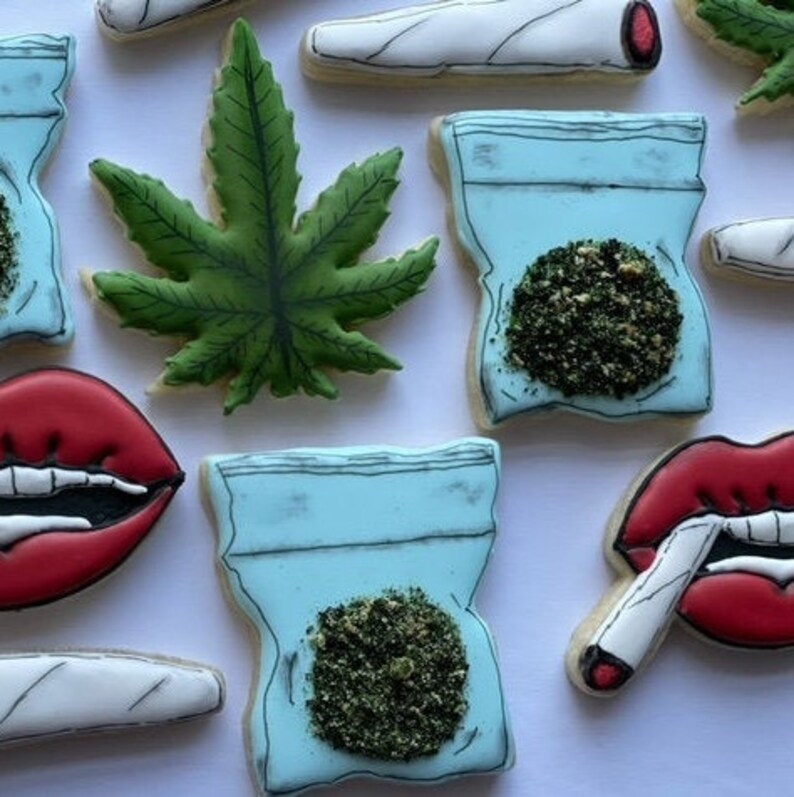 May include: Assortment of decorated sugar cookies featuring marijuana-themed designs, including a green marijuana leaf, a blue bag with green marijuana, and a red lip with a white cigarette.