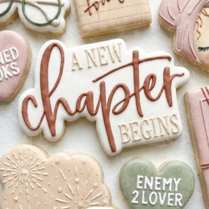 May include: A selection of decorated biscuits with a book theme. The central biscuit reads "A New Chapter Begins" in brown script. Other biscuits include glasses, a book, and a heart-shaped biscuit with the text "Enemy 2 Lover".