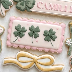Puede incluir: Una colección de galletas de azúcar decoradas. Una galleta presenta tres tréboles verdes sobre un rectángulo rosa con un borde festoneado. Otras galletas incluyen un lazo y una placa con el nombre "CAMILLE".