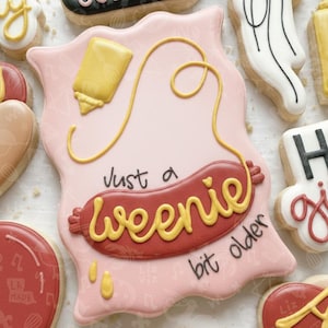 Puede incluir: Una galleta decorada con un fondo rosa y un diseño de salchicha. La salchicha es roja con mostaza amarilla y las palabras "Just a weenie bit older". Otras galletas son visibles en el fondo.