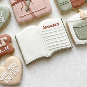 May include: A selection of decorated biscuits. One biscuit is shaped like an open book with the word "January" and a calendar grid. Other biscuits include a gift box, a heart, and a candle.