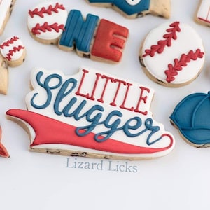 May include: Decorated sugar cookies with a baseball theme. The cookies feature baseballs, a baseball cap, the word "ONE" and the phrase "Little Slugger". The cookies are decorated in red, white and blue icing.