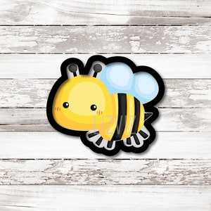 May include: A black and yellow cartoon bee cookie cutter with a light blue wing. The bee has a cute face with black eyes and pink cheeks.