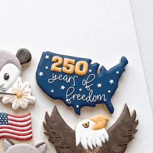 May include: Cookies decorated with patriotic themes. A blue cookie shaped like the United States has the text "250 years of freedom". Other cookies include an American flag, an eagle, and a mouse.