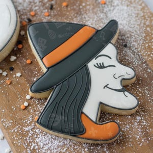 May include: A Halloween-themed cookie shaped like a witch's face, featuring a black hat with an orange band and a white face with closed eyes and a smiling mouth. The cookie is decorated with black and orange icing and sits on a wooden board.