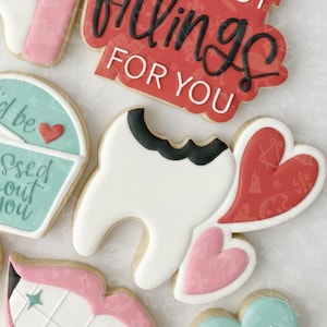 May include: Assortment of decorated sugar cookies. One cookie is shaped like a tooth with a black filling. Other cookies are heart-shaped and have the text "Fillings for You". Additional cookies are shaped like an envelope and a star.