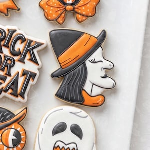 Puede incluir: Una colección de galletas de azúcar con temática de Halloween decoradas con glaseado real. Una galleta tiene forma de perfil de bruja con un sombrero negro y naranja, mientras que otra presenta el texto TRICK OR TREAT en negro y naranja.