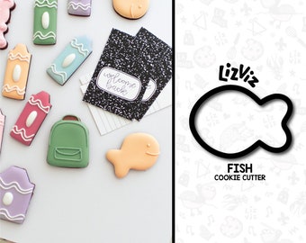 May include: A cookie cutter in the shape of a fish. The cutter is black and is on a white background with a pattern of school supplies. The text "FISH COOKIE CUTTER" is below the cutter.