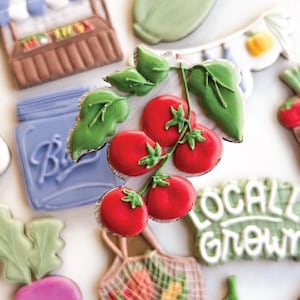 May include: An assortment of decorated cookies in the shape of fresh produce and market items. The cookies include red tomatoes, a corn cob, a farmer's market stand, and a string bag filled with vegetables. The words "LOCAL GROWN" are also visible.