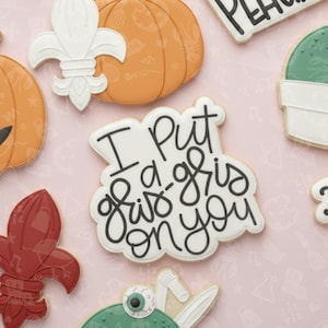 May include: A selection of decorated sugar cookies on a pink surface. Cookies include a white cookie with the text "I Put a Gris Gris on You", a pumpkin, a fleur-de-lis, and a cactus.