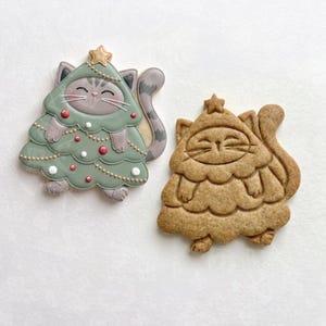 May include: Two cat-shaped cookies. One is decorated with green icing, red and white dots, and a gold star. The other is a plain, un-iced brown cookie with embossed details of a cat wearing a tree costume and a star on top.