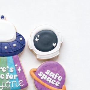 Puede incluir: Galletas de azúcar decoradas con temática espacial. Una galleta tiene forma de casco de astronauta con visera azul oscuro y detalles blancos. Otra galleta es un planeta morado con las palabras "safe space" y un anillo naranja. Una tercera galleta es un OVNI.