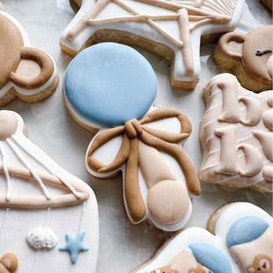 May include: Assortment of decorated sugar cookies in a baby shower theme. The cookies include a blue rattle, a baby carriage, a teddy bear, and a rocking horse, all decorated with white, brown, and blue icing.