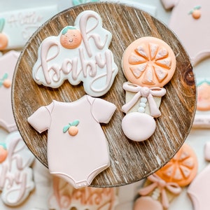 Peut inclure: Un ensemble de six biscuits d&eacute;cor&eacute;s en sucre repr&eacute;sentant une grenouill&egrave;re rose pour b&eacute;b&eacute;, un hochet et la phrase "Oh Baby" avec un joli motif orange. Les biscuits sont dispos&eacute;s sur un pr&eacute;sentoir &agrave; g&acirc;teau en bois.