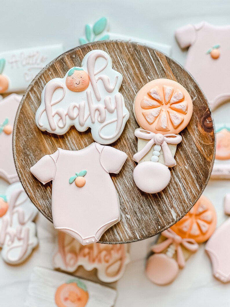 Peut inclure: Un ensemble de six biscuits d&eacute;cor&eacute;s en sucre repr&eacute;sentant une grenouill&egrave;re rose pour b&eacute;b&eacute;, un hochet et la phrase "Oh Baby" avec un joli motif orange. Les biscuits sont dispos&eacute;s sur un pr&eacute;sentoir &agrave; g&acirc;teau en bois.