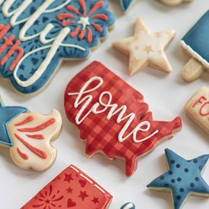 May include: Assortment of decorated sugar cookies in patriotic themes. Cookies include a red and white checkered USA-shaped cookie with the word "home", star-shaped cookies, and cookies with fireworks and "4th of July" designs.