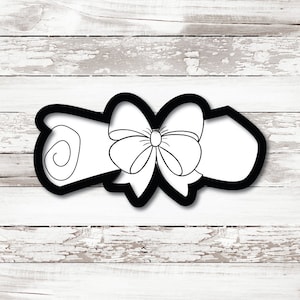 May include: A black and white cookie cutter in the shape of a rolled-up scroll tied with a bow. The scroll is white with a black outline, and the bow is detailed with white lines. The cutter is set against a white wooden background.