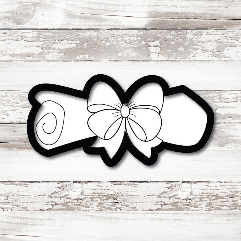 May include: A black and white cookie cutter in the shape of a rolled-up scroll tied with a bow. The scroll is white with a black outline, and the bow is detailed with white lines. The cutter is set against a white wooden background.