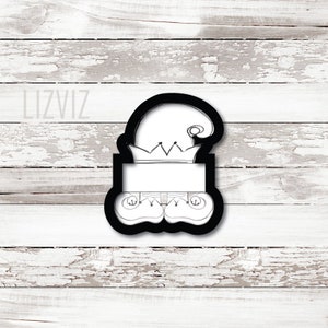 May include: Black and white cookie cutter in the shape of an elf wearing a hat and shoes.