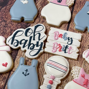 Peut inclure: Un ensemble de neuf biscuits d&eacute;cor&eacute;s pour une f&ecirc;te pr&eacute;natale. Les biscuits sont d&eacute;cor&eacute;s de gla&ccedil;age bleu et rose et pr&eacute;sentent des motifs tels que des barboteuses pour b&eacute;b&eacute;, un hochet et une pancarte qui dit "Oh Baby" et "Baby Britt".