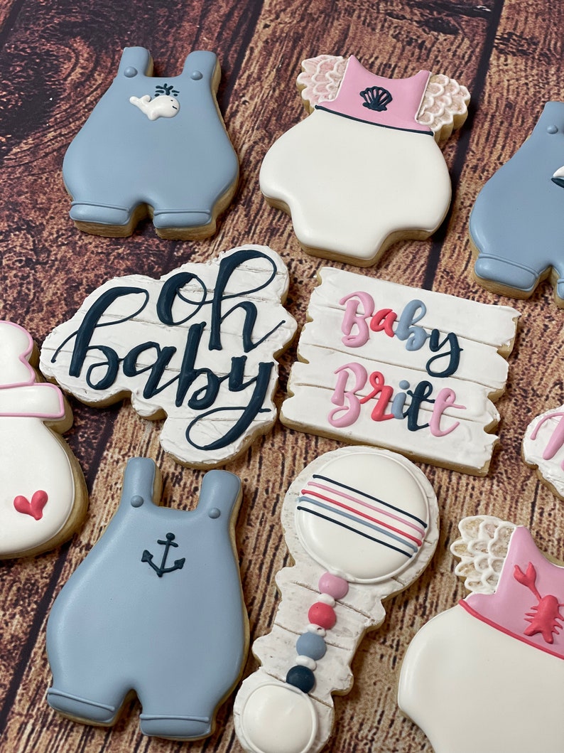 Peut inclure: Un ensemble de neuf biscuits d&eacute;cor&eacute;s pour une f&ecirc;te pr&eacute;natale. Les biscuits sont d&eacute;cor&eacute;s de gla&ccedil;age bleu et rose et pr&eacute;sentent des motifs tels que des barboteuses pour b&eacute;b&eacute;, un hochet et une pancarte qui dit "Oh Baby" et "Baby Britt".