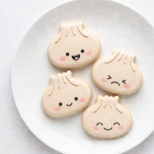May include: Four beige, dumpling-shaped sugar cookies arranged on a white plate. Each cookie features a hand-piped face with black eyes, a mouth, and pink blush. Three cookies display smiling expressions, while one cookie displays a frowning expression.