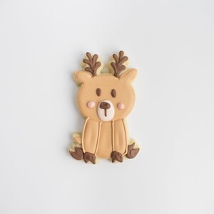 May include: A reindeer-shaped biscuit with a light brown base. It has a tan body, brown antlers, and dark brown hooves. The biscuit is decorated with pink cheeks, a white nose, and brown eyes. The design is detailed and festive.