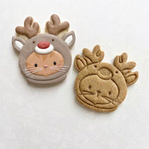 May include: Two reindeer-shaped cookies. One is decorated with brown and tan icing, featuring a cat face, a red nose, and white whiskers. The second cookie is an uniced, plain brown gingerbread reindeer with embossed facial details.