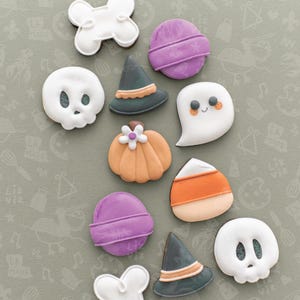 May include: A collection of Halloween-themed cookies, including a skull, ghost, pumpkin, witch hat, bone, and candy corn. The cookies are decorated with white, orange, purple, and green icing. The cookies are arranged on a light green surface.