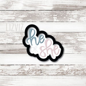 May include: A black and white cookie cutter in the shape of a cloud with the words "he or she" written in blue and pink.