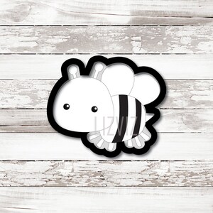 May include: Black and white cookie cutter in the shape of a cartoon bee. The bee has a round head with two small eyes and two antennae. The body is striped black and white.