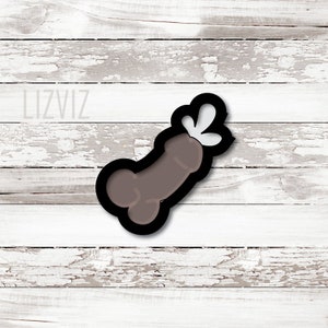 May include: A brown cookie cutter in the shape of a penis with a black outline and white accents. The cookie cutter is on a white wooden background. The text "LIZVIZ" is in the upper left corner.