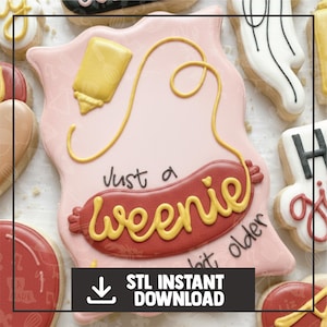 Op de afbeelding: Een roze glazuurkoekje in de vorm van een hotdog met gele mosterd en de woorden "Just a Weenie, a bit older" in gele glazuur. Het koekje heeft de vorm van een rode hotdog en is omringd door andere versierde koekjes.