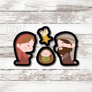 May include: A set of three Christmas-themed cookie cutters featuring a nativity scene. The set includes Mary, Joseph, baby Jesus, and a star, all with black outlines and cartoon-style illustrations. The background is a white wooden surface.