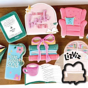 May include: A set of nine cookie cutters featuring book-themed designs. The cutters are made of metal and have a silver finish. The designs include a stack of books, a book with a bow, a book with the text "in my book era", a book with the text "Chapter Two", a book with the text "Read Banned Books", a cup of coffee, an open book, a bookshelf, and an armchair.