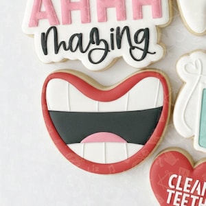 May include: Decorated sugar cookies with a dental theme. One cookie is shaped like a smiling mouth with red lips, white teeth, and a black interior. Other cookies spell "AHHHmazing" and "CLEAN TEETH".