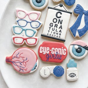 May include: A collection of eye-themed sugar cookies including four pairs of glasses in pink, blue, teal, and red, a white eye chart with the text CONGRATULATIONS DOCTOR, a blue bow, an eyeball, a red plaque with eye-conic girlie, a contact lens case, and eye drops.