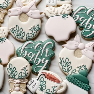 Peut inclure: Un ensemble de neuf biscuits d&eacute;cor&eacute;s sur le th&egrave;me du b&eacute;b&eacute;. Les biscuits sont d&eacute;cor&eacute;s de feuilles vertes, de n&oelig;uds roses et de gla&ccedil;age blanc. Les biscuits pr&eacute;sentent divers motifs li&eacute;s au b&eacute;b&eacute;, notamment un hochet, une grenouill&egrave;re, une tasse de th&eacute; et un biberon. Les biscuits sont d&eacute;cor&eacute;s des mots "oh baby" et "hello baby".