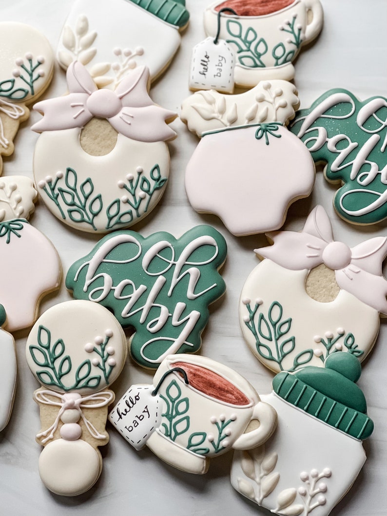 Peut inclure: Un ensemble de neuf biscuits d&eacute;cor&eacute;s sur le th&egrave;me du b&eacute;b&eacute;. Les biscuits sont d&eacute;cor&eacute;s de feuilles vertes, de n&oelig;uds roses et de gla&ccedil;age blanc. Les biscuits pr&eacute;sentent divers motifs li&eacute;s au b&eacute;b&eacute;, notamment un hochet, une grenouill&egrave;re, une tasse de th&eacute; et un biberon. Les biscuits sont d&eacute;cor&eacute;s des mots "oh baby" et "hello baby".
