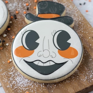 May include: A Halloween-themed sugar cookie decorated with a smiling face design. The cookie features a white base with black and orange accents, including a top hat. The face has large eyes, rosy cheeks, and a wide grin. Sprinkles and powdered sugar are scattered around the cookie.